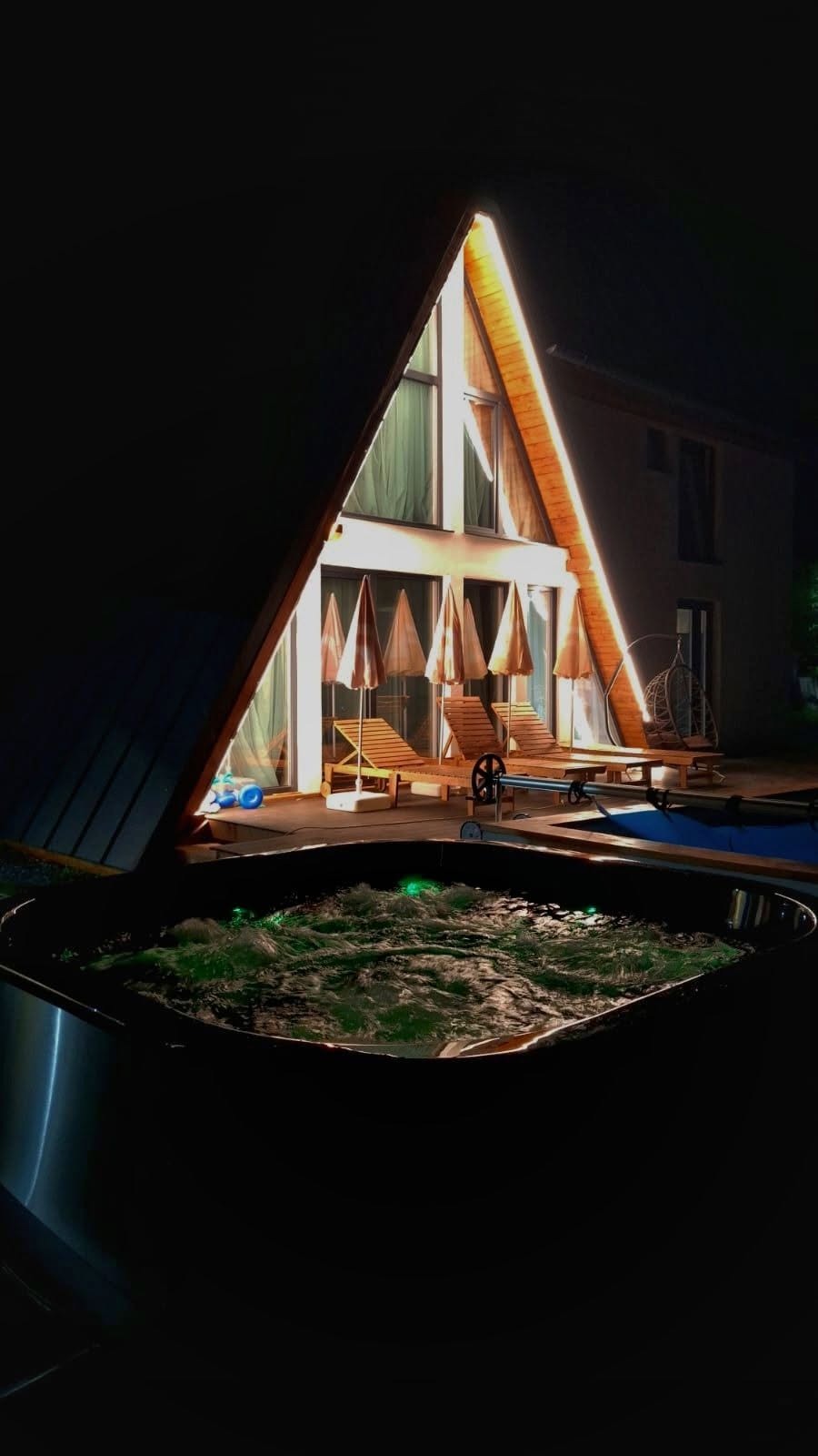 Căsuța A-frame luminată noaptea, cu piscina în față